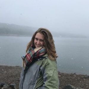 Carlee is in front of a lake with fog in the background and a rocky shore, wearing a big scarf and raincoat
