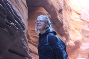 Eliza is wearing glasses, a raincoat and backpack, smiling in front of a sandstone canyon in the background