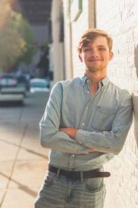 Jacob is on a sidewalk in an urban setting, leaning against a building with arms crossed smiling