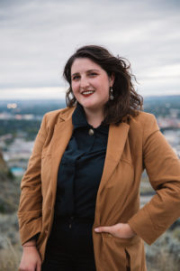 Julia is wearing a blazer jacket and standing at a vista point overlooking a city. She has shoulder length dark hair and is smiling with her hand in her pocket