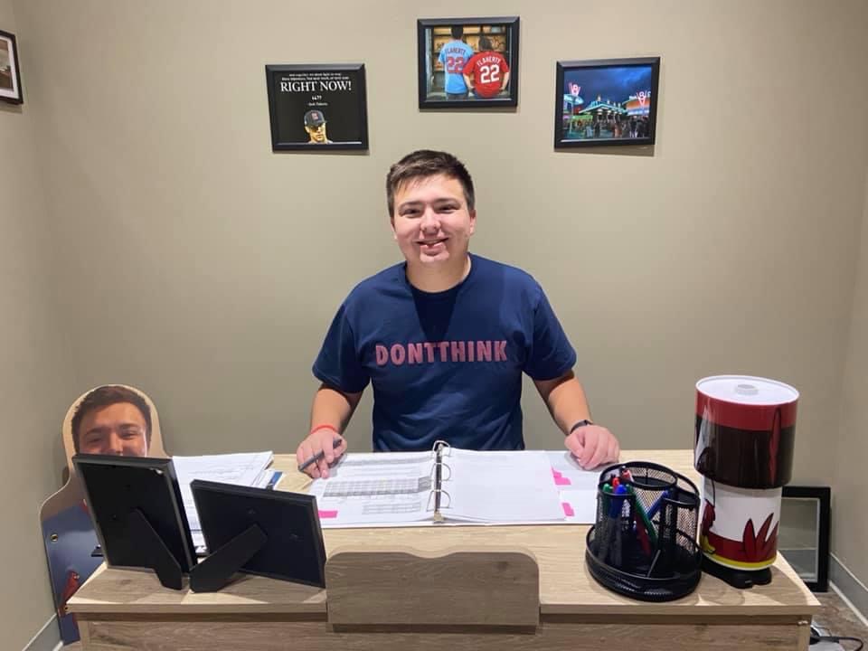Keaton is sitting at a desk, with several framed pictures behind them and documents on the desk, and is smiling with their hands resting on the desk