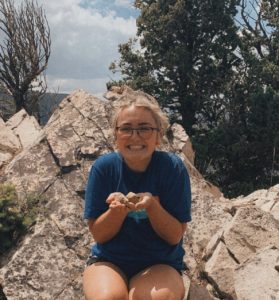 Lauryn is sitting on large rocks with trees behind her, and holding a small rock in her hands, smiling. She has her hair in a bun and is wearing glasses.