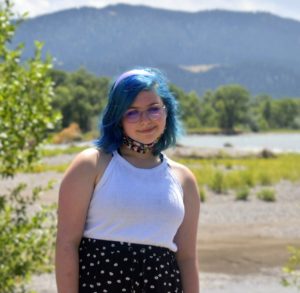 Marina has short blue hair and is standing in front of a wide river bed with mountains in the far distance