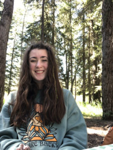 Scout has long dark hair and is smiling in a forest area with tall trees behind her
