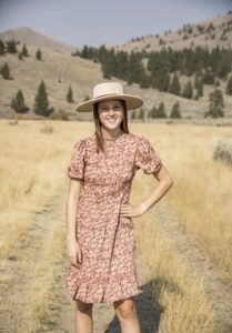 Frances is standing in front of a wheat field, wearing a wide brim hat and sundress